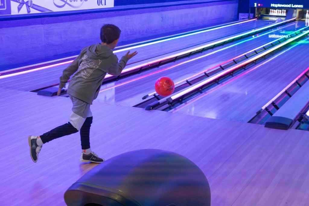 A child bowling in a brightly lit bowling alley, launching a red bowling ball down the lane with colorful lights reflecting on the polished wooden floor.