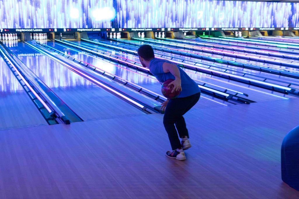 A person in a blue shirt preparing to bowl in a brightly lit bowling alley with colorful LED lights and multiple lanes.