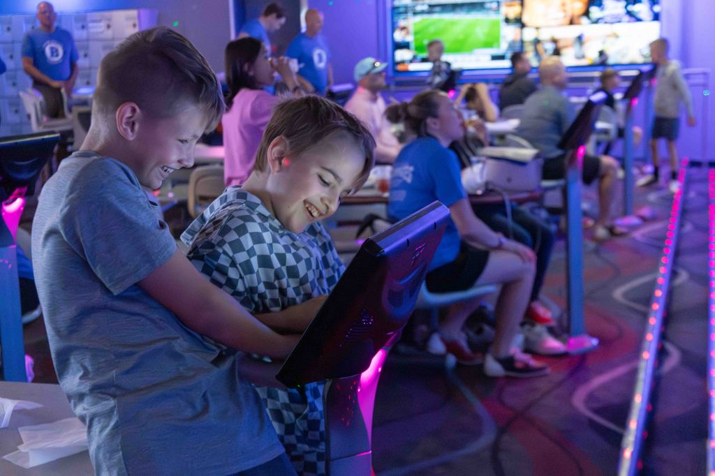 Two boys laughing while using a touchscreen gaming console in a colorful arcade environment, with other people enjoying various activities in the background.