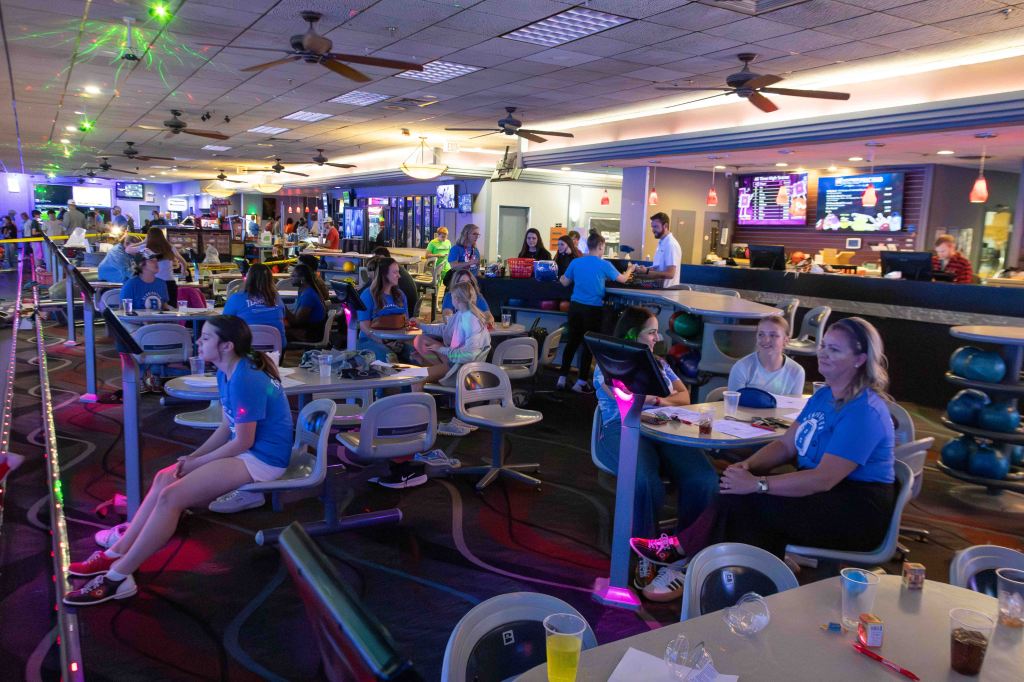 A vibrant bowling alley scene with people seated at tables, some wearing blue shirts, and a bar area in the background. Colorful lights and screens are visible, creating a lively atmosphere.