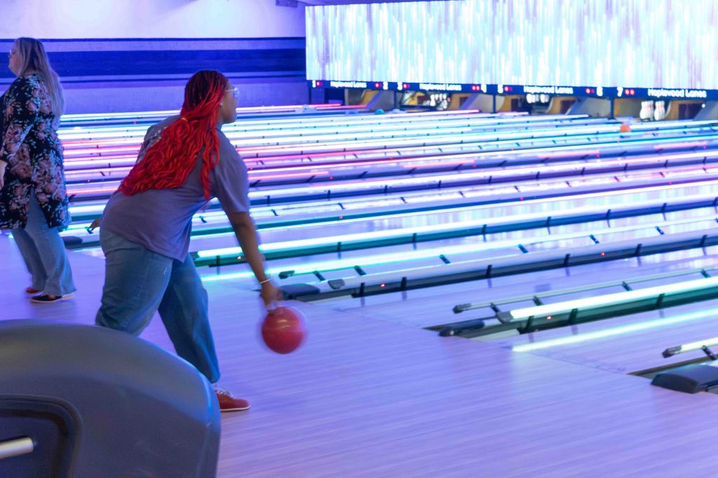 A person with long red hair is preparing to bowl at a bowling alley, while another person walks in the background. The bowling lanes are brightly lit with colorful lights.