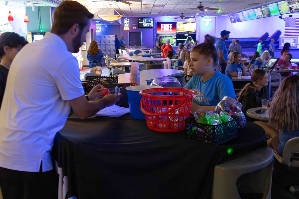A staff member interacts with a young girl at a registration table filled with colorful items in a vibrant bowling alley, with people enjoying activities in the background.