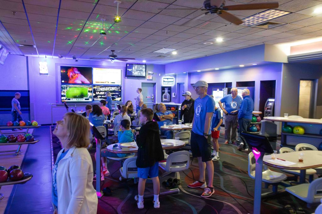 A lively bowling alley scene featuring a diverse group of people, including children and adults, enjoying their time. Colorful bowling balls are visible on the racks, and screens display scores and entertainment. The interior is decorated with purple lighting, creating a fun atmosphere.