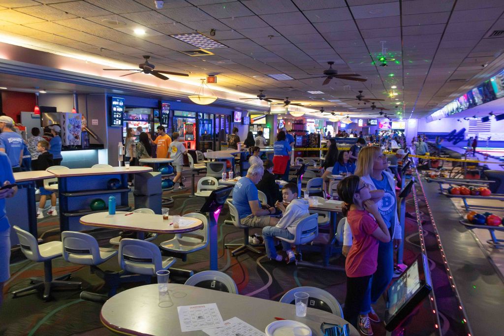 A crowded bowling alley with people of all ages enjoying games and activities. The scene includes tables with drinks, bowling balls, and colorful lighting in the background.