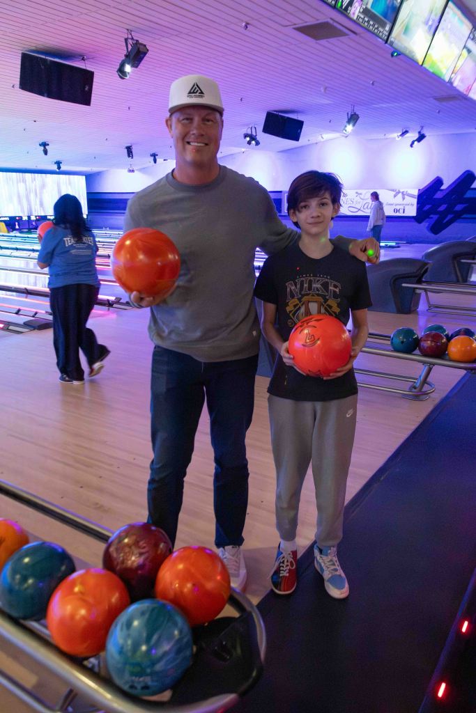 A smiling man and a boy holding bowling balls, standing in a bowling alley with colorful bowling balls in the foreground and bowling lanes in the background.