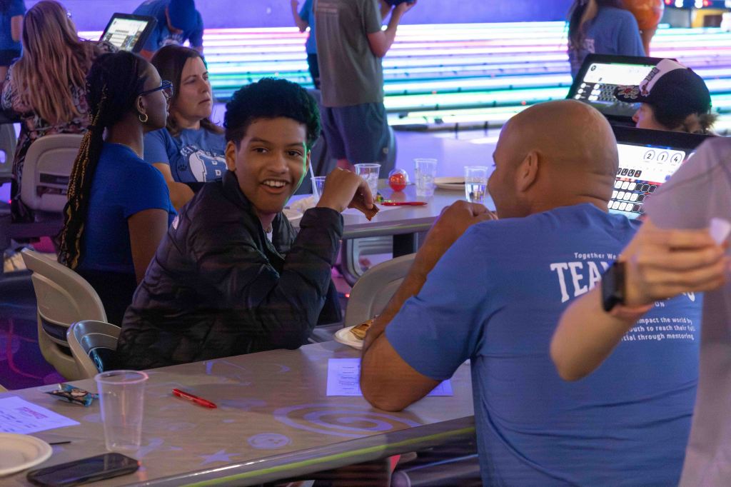 Two individuals smiling and talking at a bowling event, with others in the background. One is wearing a black jacket, and the other is wearing a blue shirt that says 'TEAM'.
