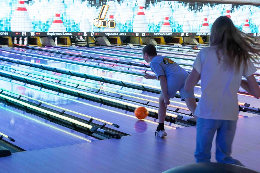 A child bowling on a lane at a bowling alley, with a bright orange bowling ball in motion and colorful neon lights illuminating the surroundings.