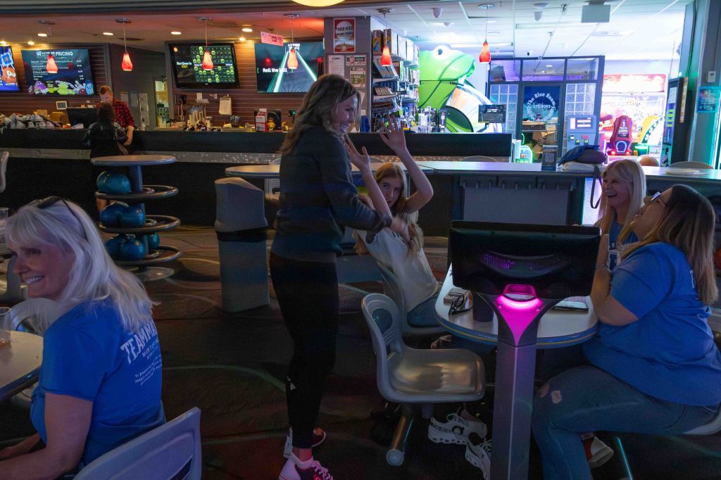 A lively bowling alley scene with several people interacting and enjoying themselves, featuring a bar area in the background and bowling balls visible.