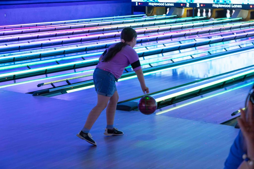 A child in a pink shirt and denim shorts is bowling at a bowling alley with colorful neon lights.