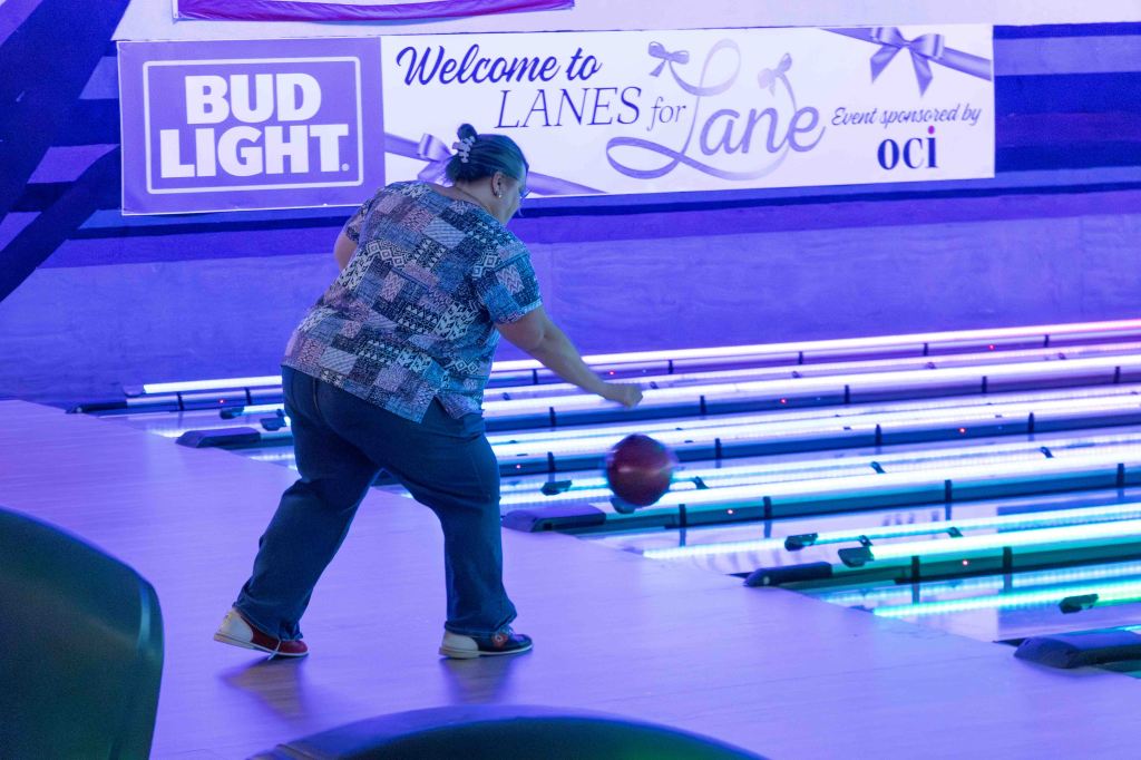 A woman bowling in a brightly lit bowling alley, with colorful lane lights and a welcome sign for an event.
