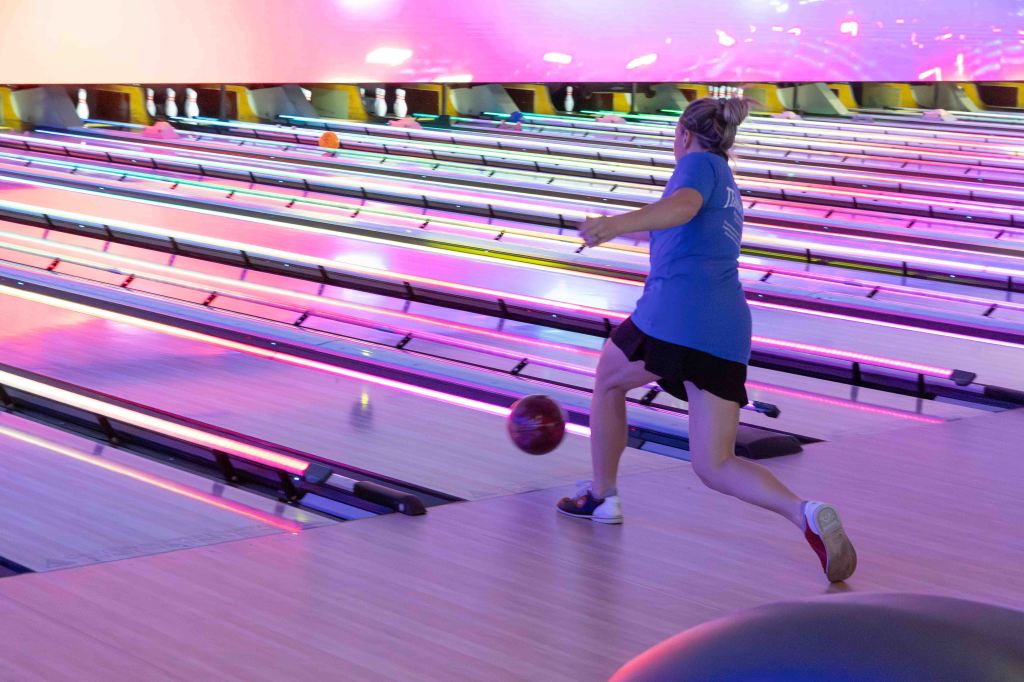 A person throwing a bowling ball down a brightly lit bowling lane with colorful lights and pins in the background.