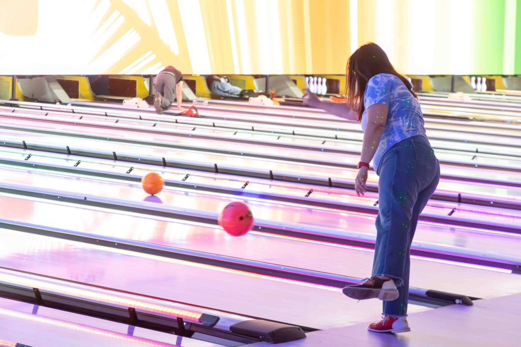 A woman bowling at a vibrant bowling alley with neon lights and two bowling balls in motion.