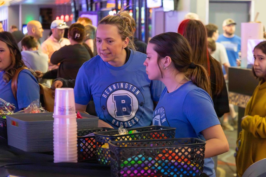 Two women in blue shirts engage at a booth, surrounded by baskets filled with colorful items, with a crowd visible in the background.