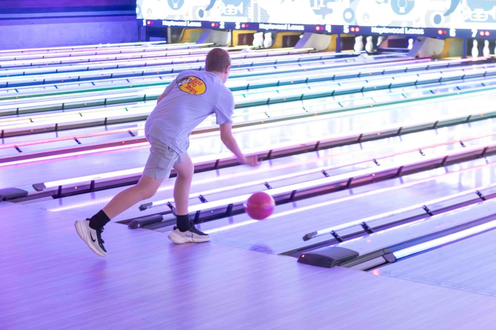 A child in a light blue shirt bowls a bright orange bowling ball down a brightly lit alley with colorful neon lights.