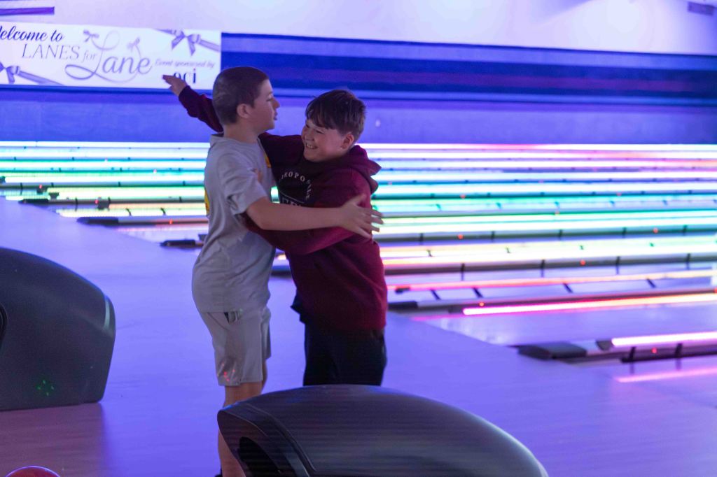 Two boys happily hugging at a bowling alley with illuminated lanes in the background.