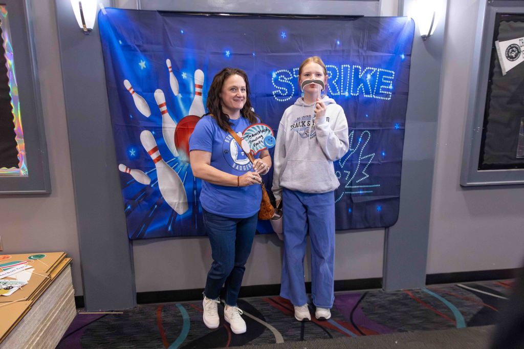 Two women posing together in front of a bowling-themed backdrop, smiling and holding props. The backdrop features bowling pins and a stylized 'Strike' sign.