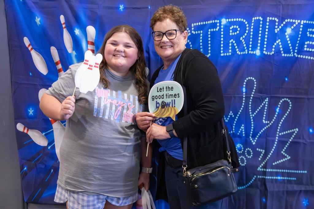 Two smiling women posing together at a bowling-themed event, holding props shaped like bowling pins and a sign that says 'Let the good times bowl!' with a festive blue backdrop.