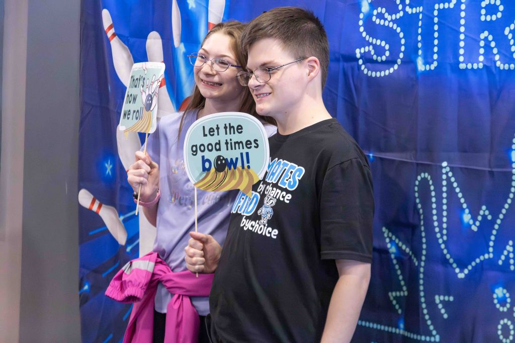 Two individuals pose for a photo holding fun signs that say 'That's how we roll!' and 'Let the good times bowl!!', in front of a bowling-themed backdrop.