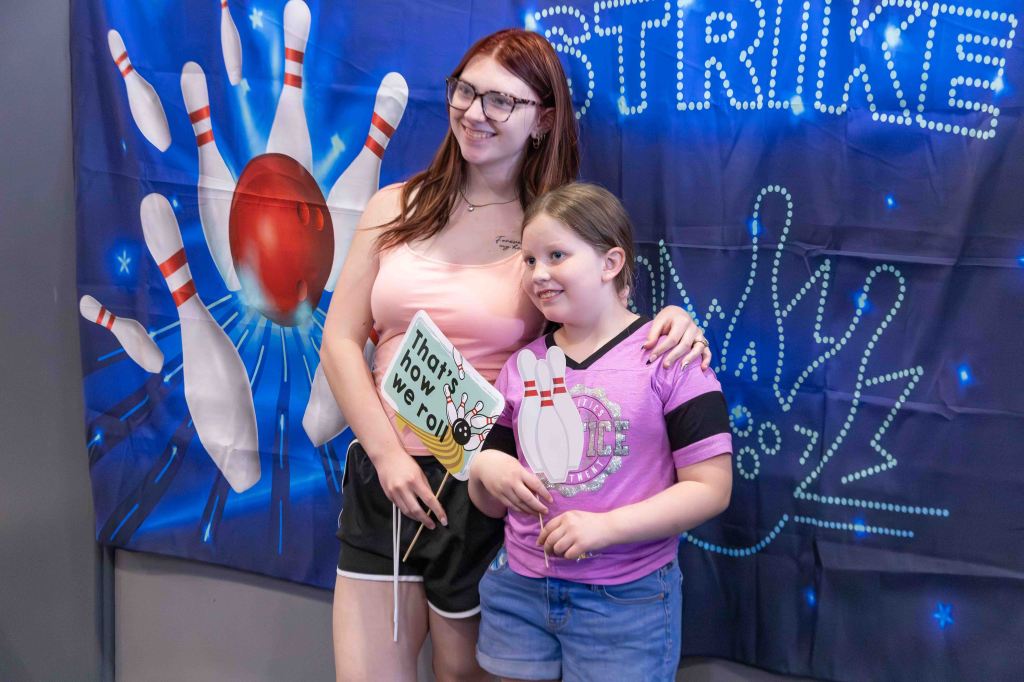 Two young girls posing together with a bowling-themed backdrop, one holding a sign that says 'That's how we roll'.