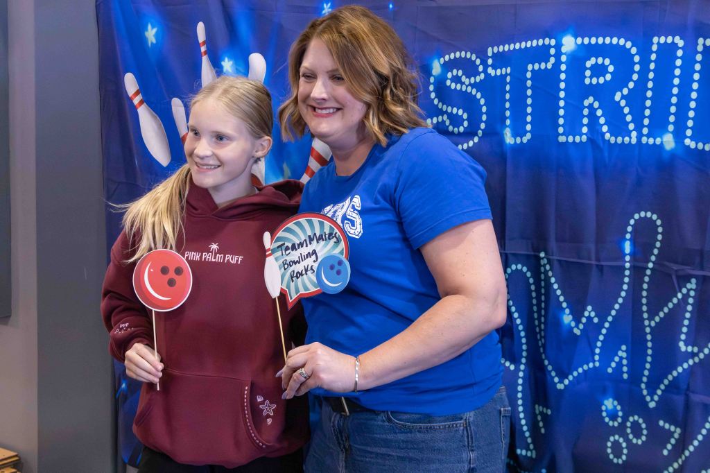 Two smiling women pose together with colorful props at a bowling-themed event, featuring a backdrop with bowling pins and lights.