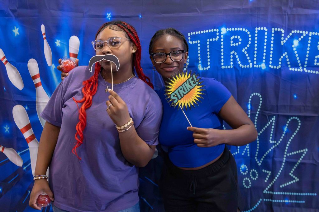 Two women pose with props for a photo booth against a bowling-themed backdrop, one holding a fake mustache and the other a 'Strike!' sign.