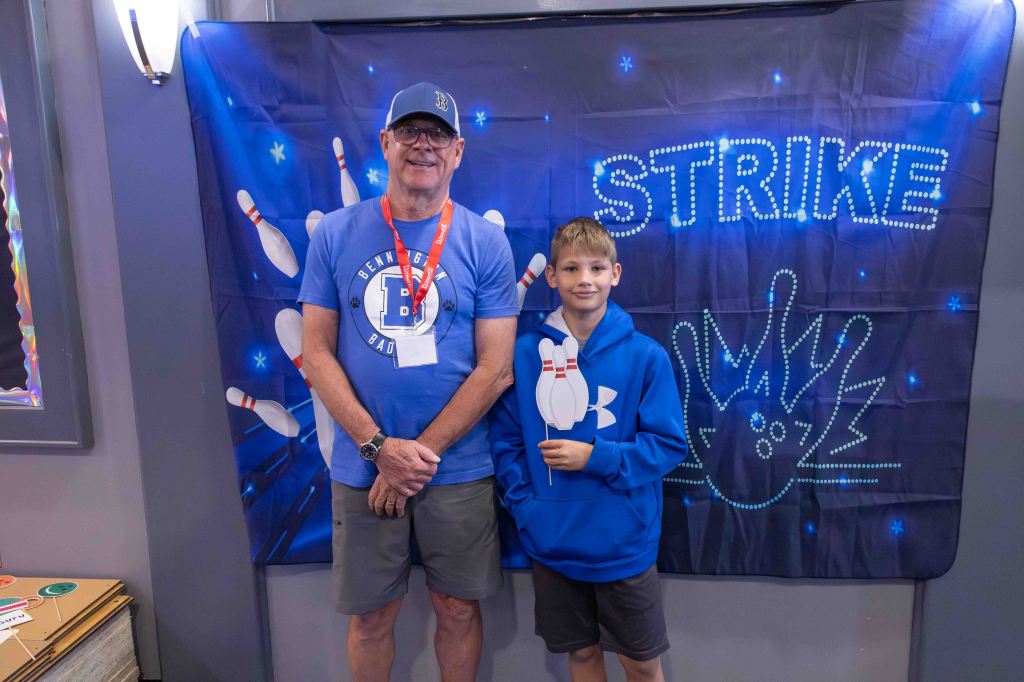 A smiling man and a young boy standing in front of a colorful bowling-themed backdrop with the word 'STRIKE' and bowling pins.