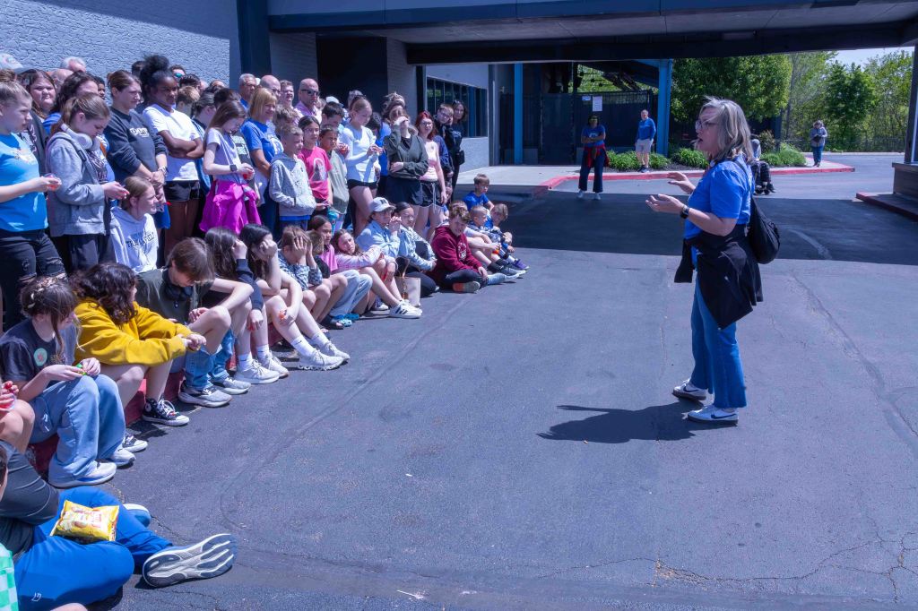 A group of children and teenagers sitting on the ground attentively listening to a woman speaking in front of them, outdoors in a sunny environment.