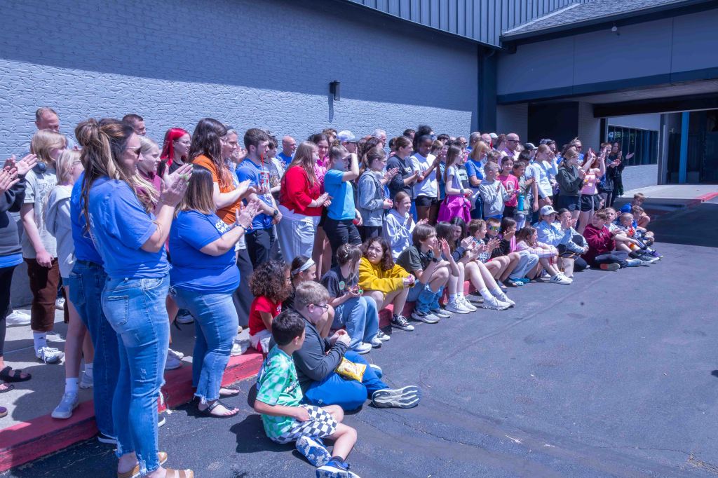 A large group of people, including children and adults, are gathered outdoors, clapping and enjoying a moment together. They are standing and sitting on a curb near a building, with a variety of colorful clothing visible.