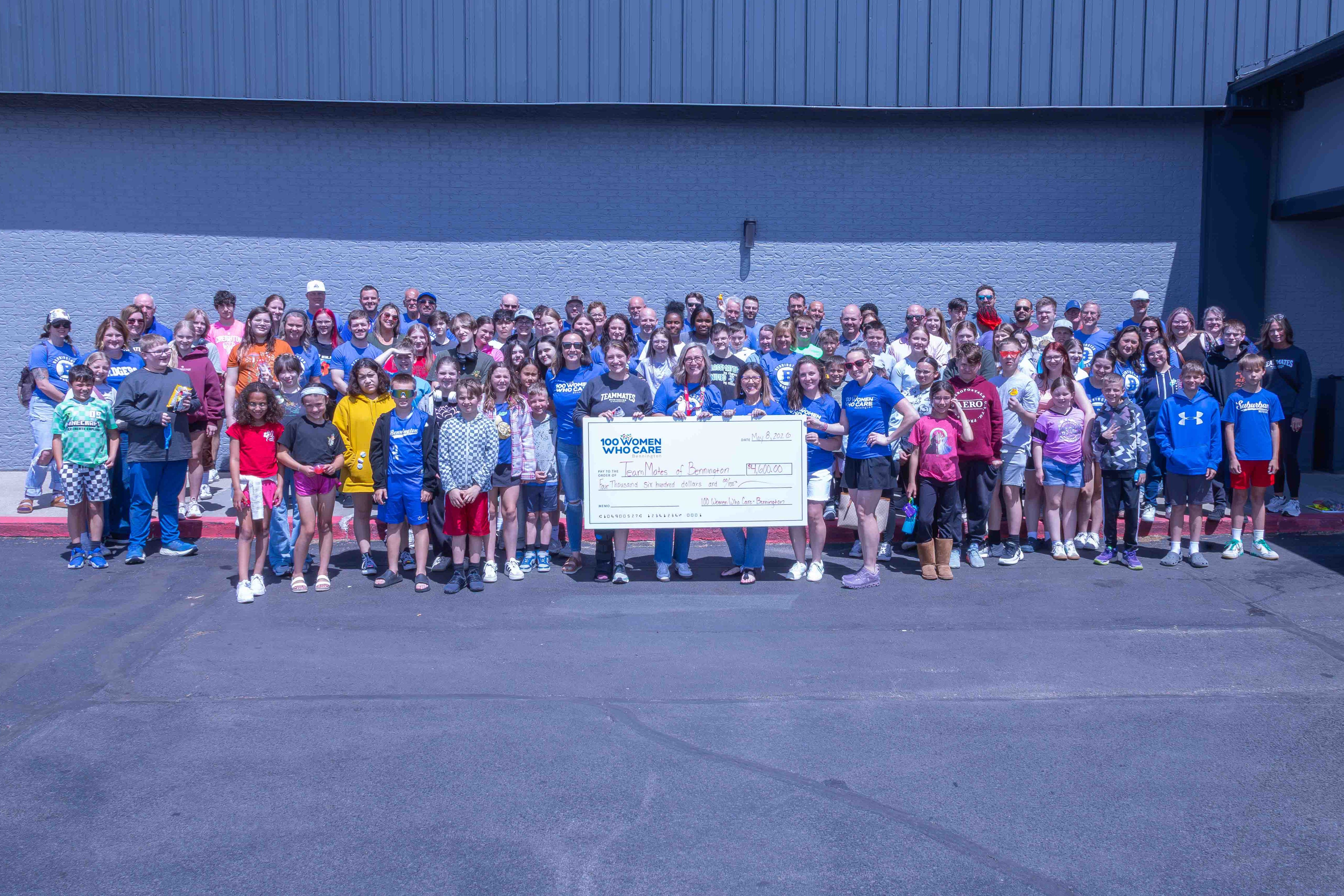 A large group of people, including children and adults, gathered outdoors in front of a building, smiling and holding a large check. The event appears to be a celebration or charity fundraising initiative.