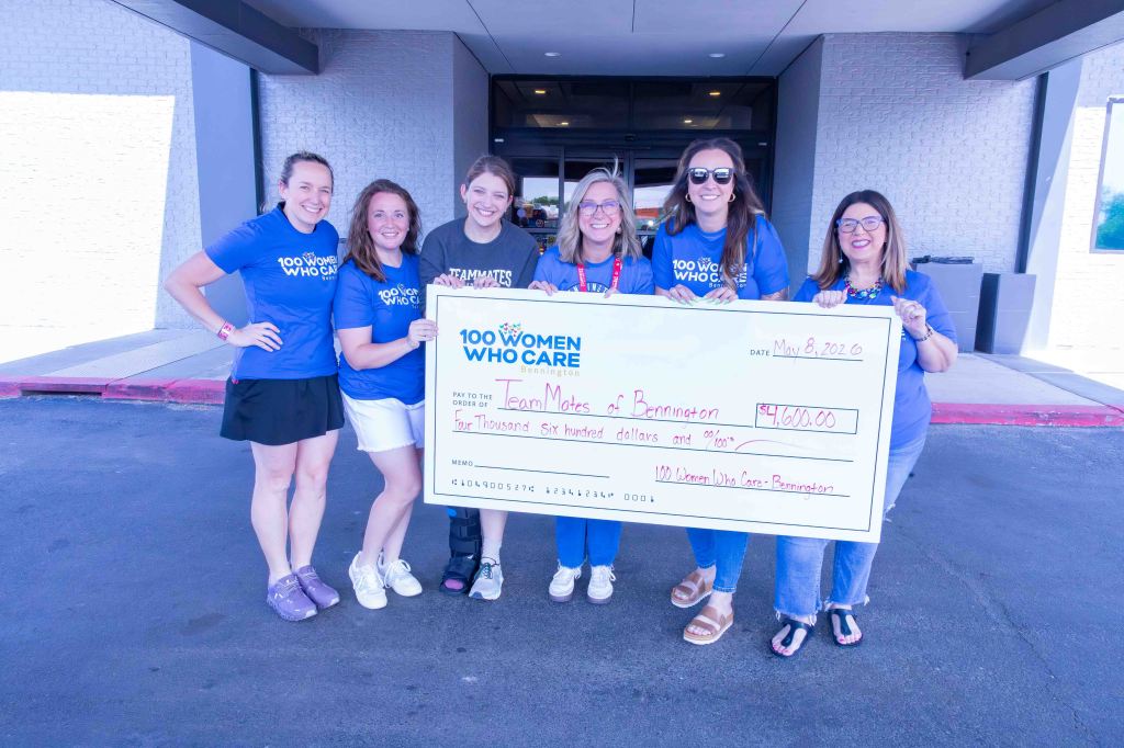 A group of women holding a large check for $4,600, presented to 'Team Moes of Bennington' on May 8, 2016, outside a building. All are wearing blue '100 Women Who Care' t-shirts.