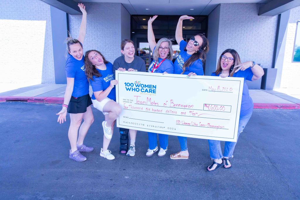 A group of six women celebrating outside a building, holding a large check for $4,600 made out to 'Team Mates of Bennington' as part of the '100 Women Who Care' initiative, with joyful expressions and poses.