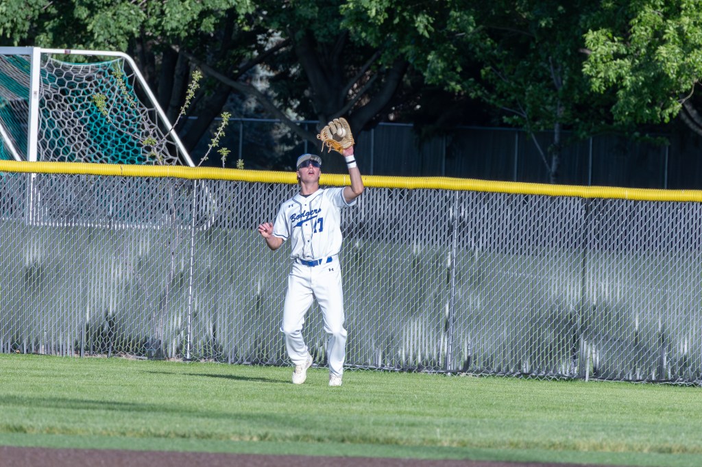 A baseball player in white and blue uniform is standing in the outfield, preparing to catch a ball. The player is wearing a cap and sunglasses, with a baseball glove raised, and the background features a chain-link fence and trees.