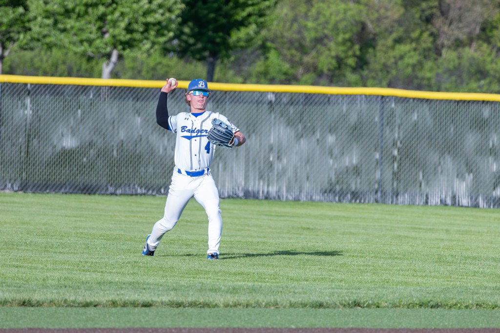 A baseball player in a white and blue uniform stands in an outfield position, preparing to throw a baseball. He wears a blue cap and has a focused expression.