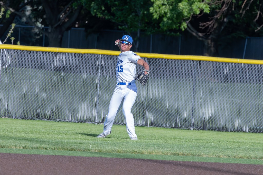 A baseball player wearing a white uniform and a blue hat is standing in the outfield, preparing to catch a ball.