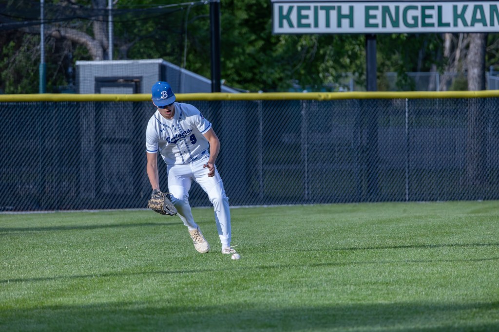A baseball player in a white uniform and blue cap is fielding a ball in the outfield, with a green fence and a sign in the background.
