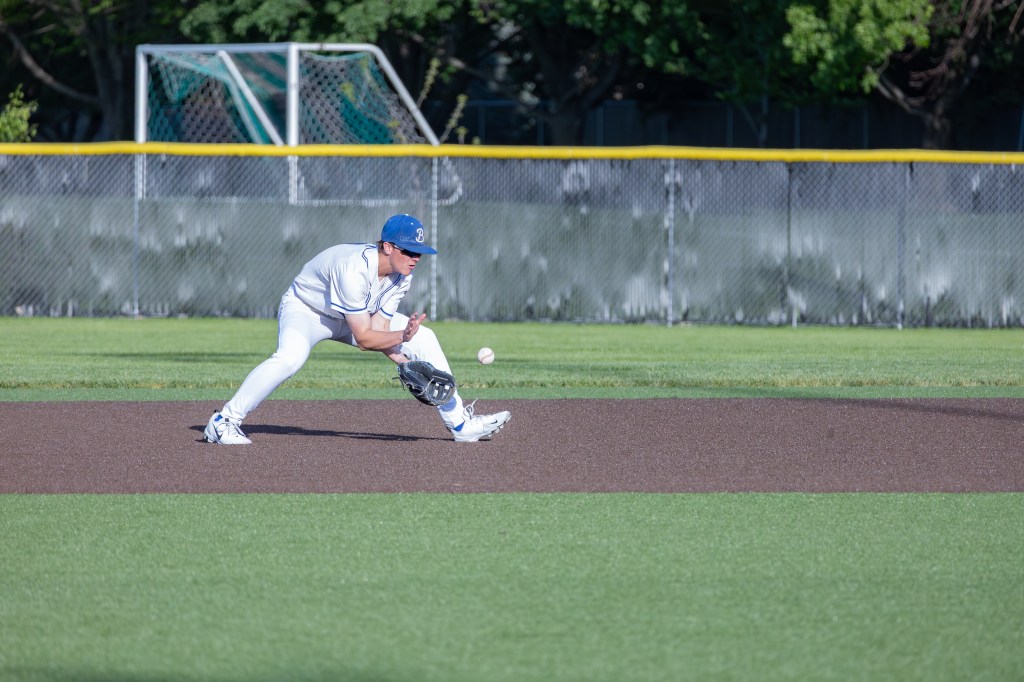 A baseball player in a white uniform and blue cap fielding a ground ball on a grassy baseball field.