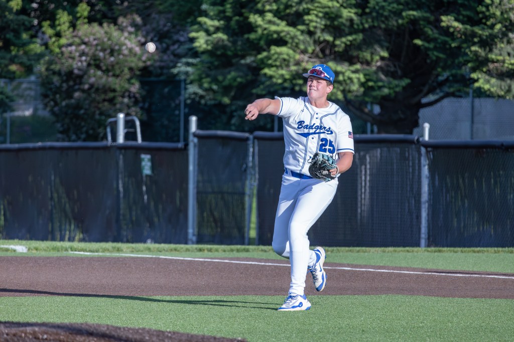 A baseball player wearing a white jersey and blue cap throws a baseball while standing on a field.