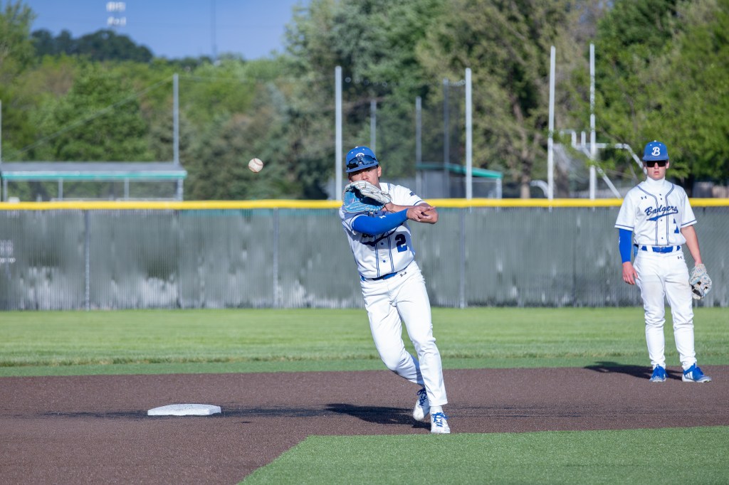 A baseball player in a blue and white uniform throwing a baseball from the infield, with another player in the background observing.