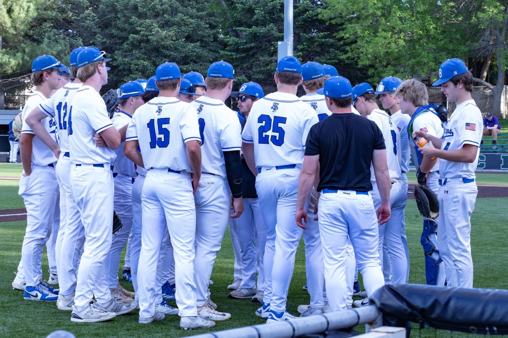 A baseball team in white uniforms with blue hats huddled together on the field, discussing strategies.