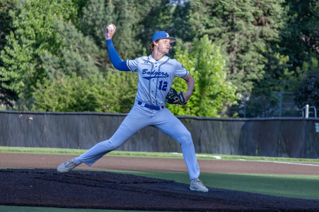 A baseball player in a blue and white uniform is on the pitcher's mound, in a dynamic throwing position, with trees in the background.