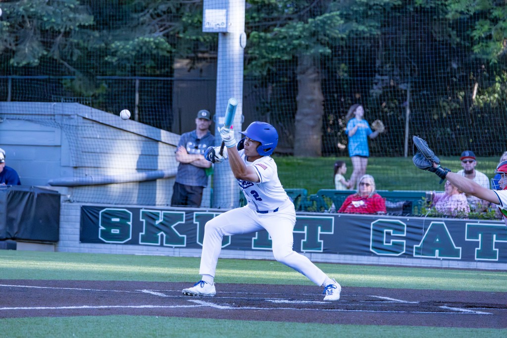A young baseball player in a blue helmet and white uniform swings at a pitched ball during a game, with spectators in the background.