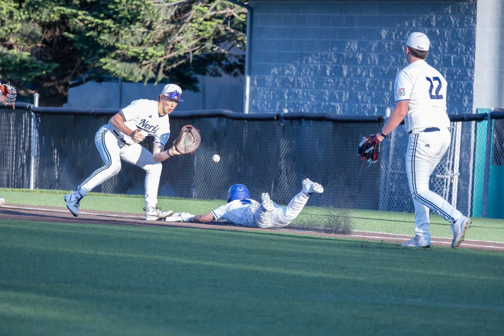 A baseball player dives for a catch as another player reaches for a throw. The scene captures the action on a baseball field with players in uniforms, demonstrating athleticism and teamwork.