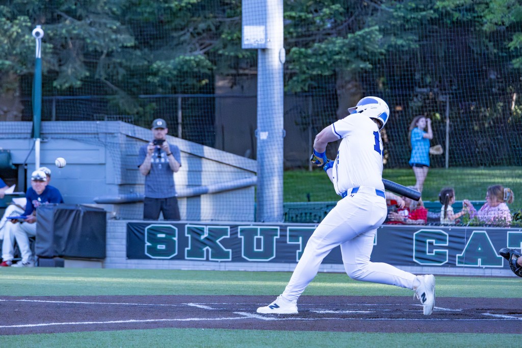 A baseball player in a white uniform swings at a pitch during a game, with spectators in the background.