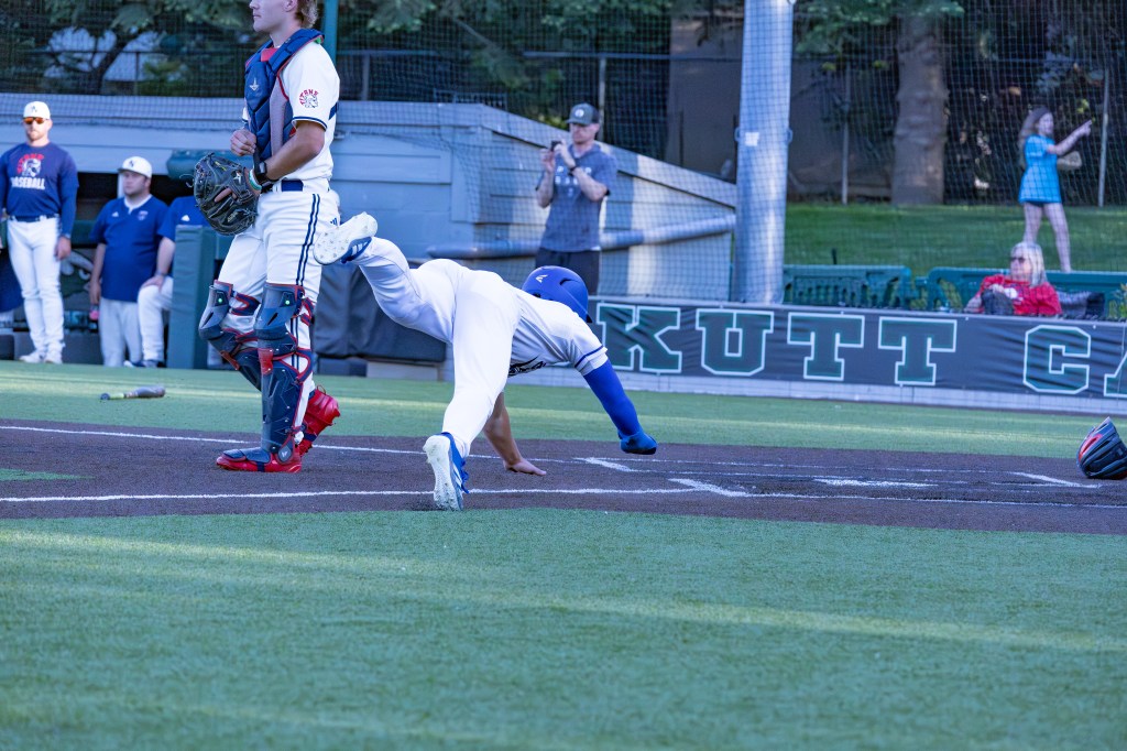 A baseball player sliding into home plate during a game, with a catcher in position to receive the play. Other players are visible in the background.