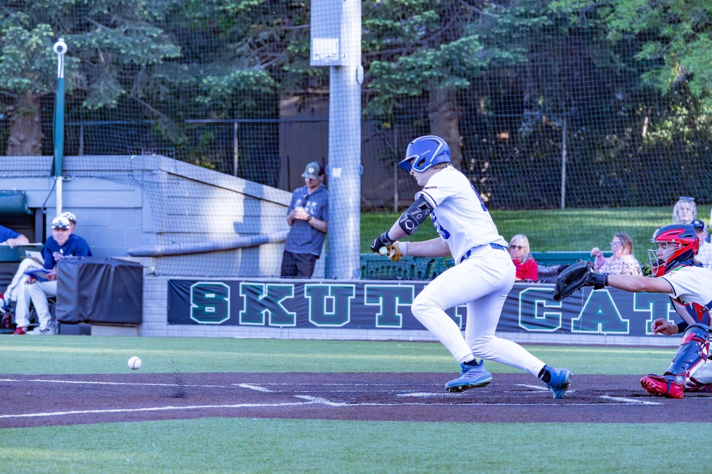 A baseball player in a white uniform is poised to hit a ball that is nearing the plate, while a catcher in red gear prepares to receive the pitch. In the background, teammates and spectators are visible.