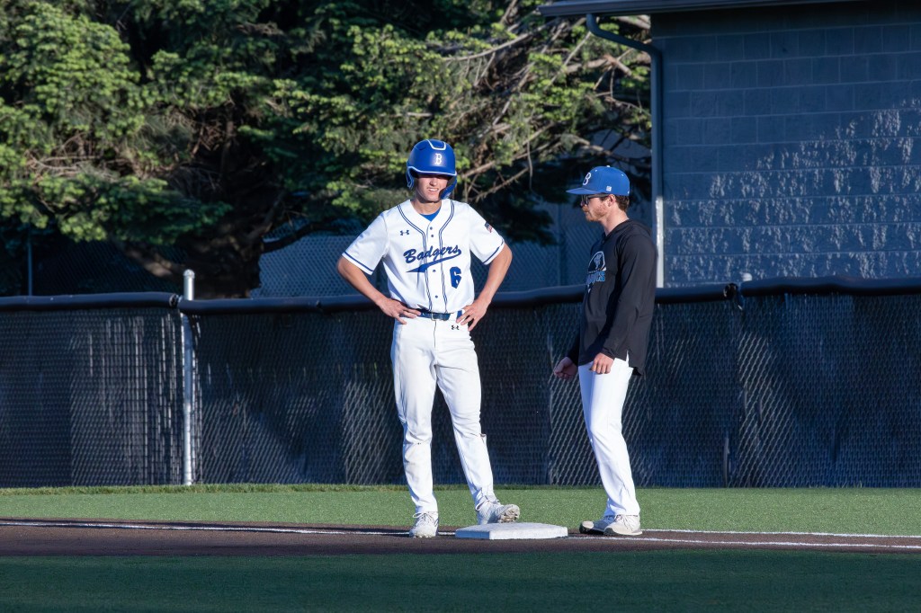 Two baseball players on the field; one standing on first base while the other, in a black hoodie, talks to him.
