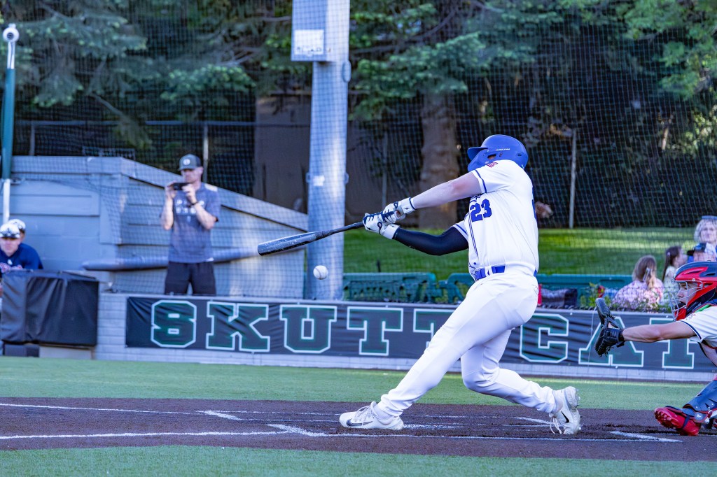 A baseball player in a white uniform swings at a pitch during a game, while a catcher in red gear crouches behind home plate. Spectators can be seen in the background.