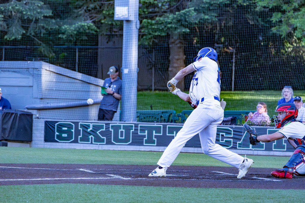 A baseball player in a white uniform swings at a pitch during a game, with the ball just leaving the pitcher's hand. A catcher in red is crouched behind home plate, and spectators are visible in the background.