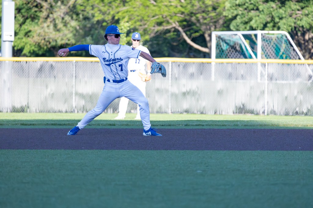 A baseball player in a white and blue uniform is about to throw a baseball from the infield, while another player stands in the background.