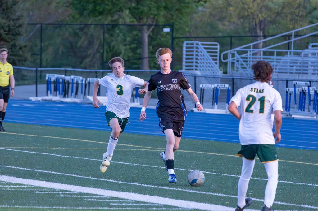 A soccer player in a black uniform dribbles the ball down the field, pursued by two opponents in white and green uniforms.